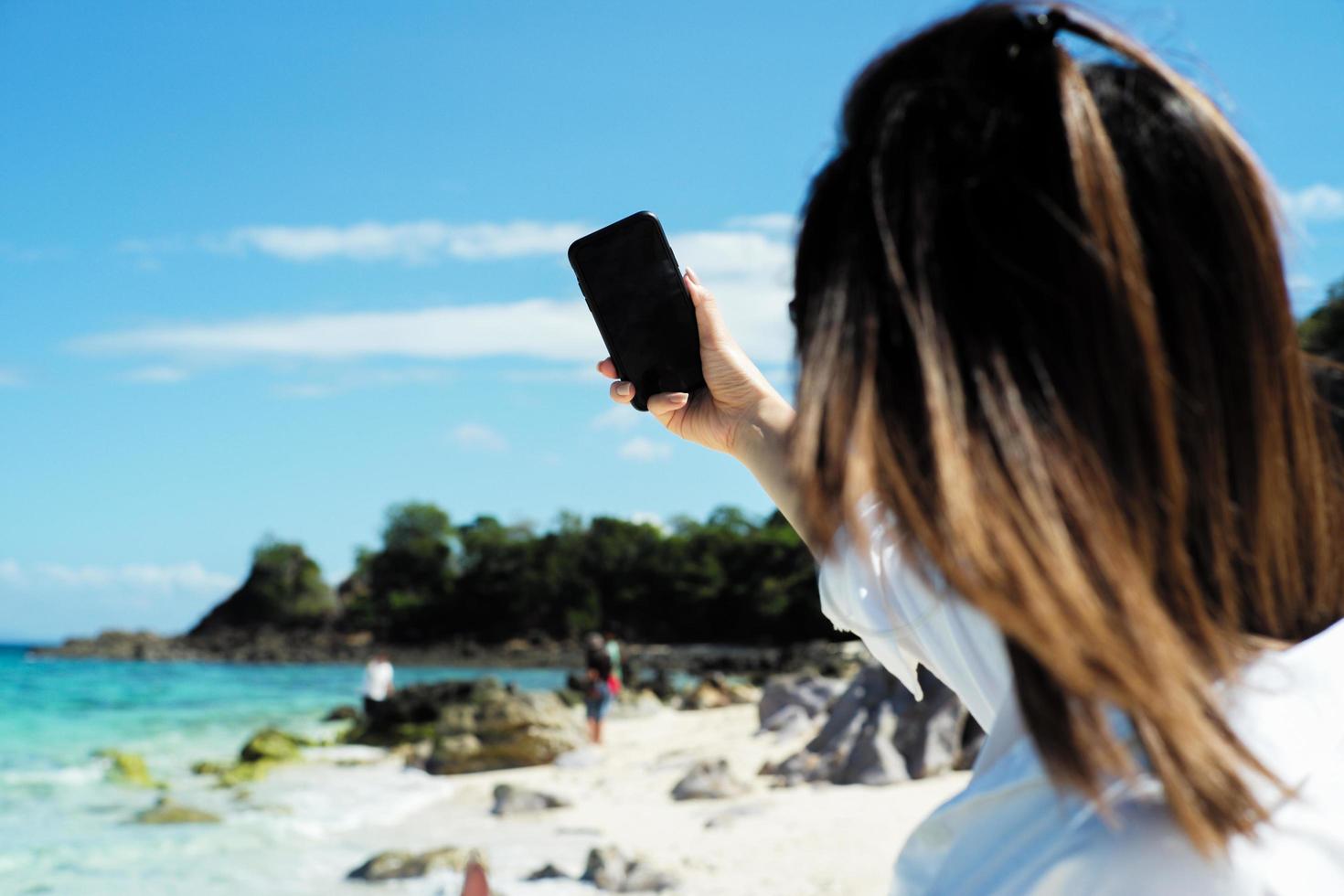 Back portrait of female tourist holding a mobile phone and taking a selfie picture with a blurred seascape and people in the background. Selective focus on blank screen of mobile held by the woman for a selfie. photo