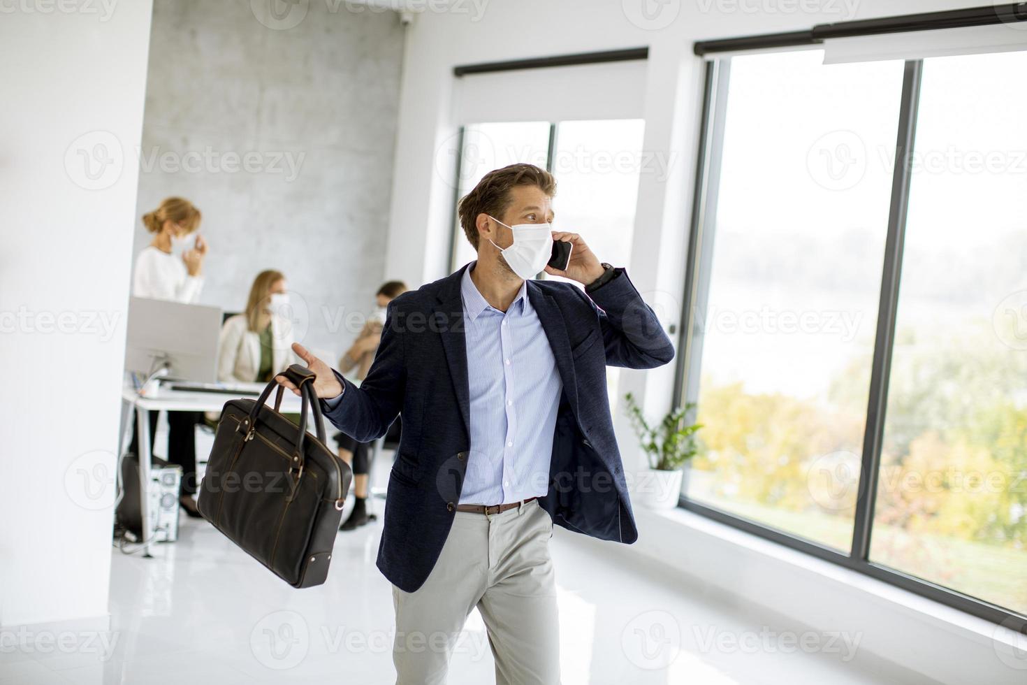 Masked businessman taking on phone with briefcase photo