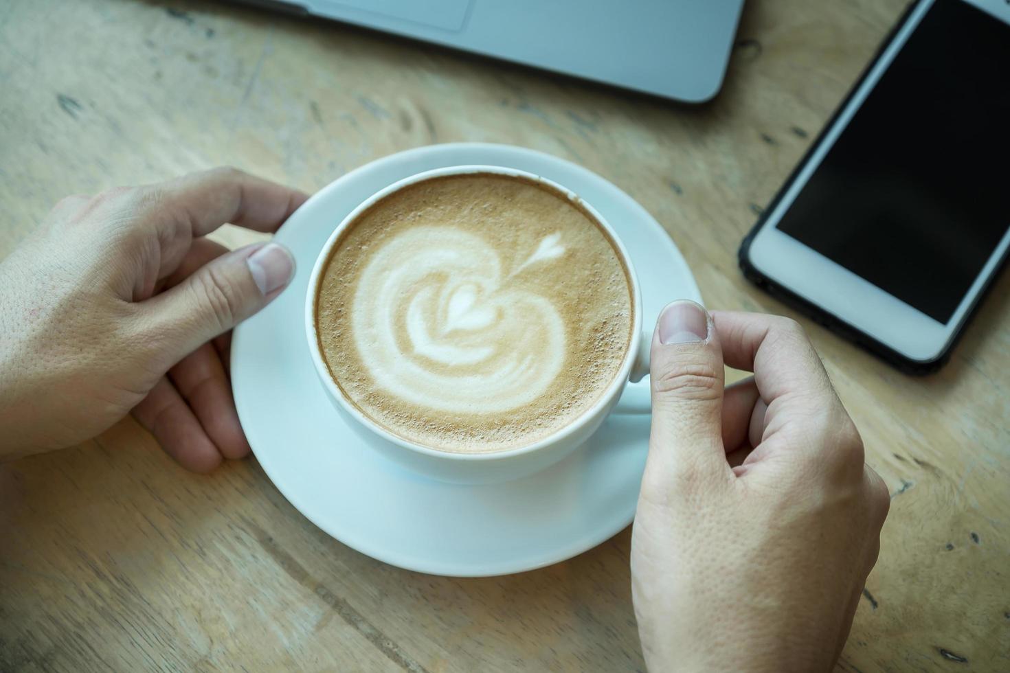 A man hand holding coffee cup on wood desk with smartphone and laptop computer in office photo