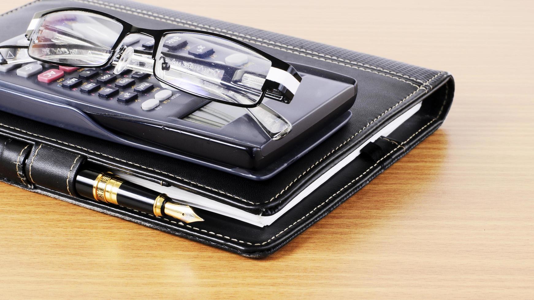 Business book and fountain pen with calculator and eyeglasses on wooden desk for work place concept photo