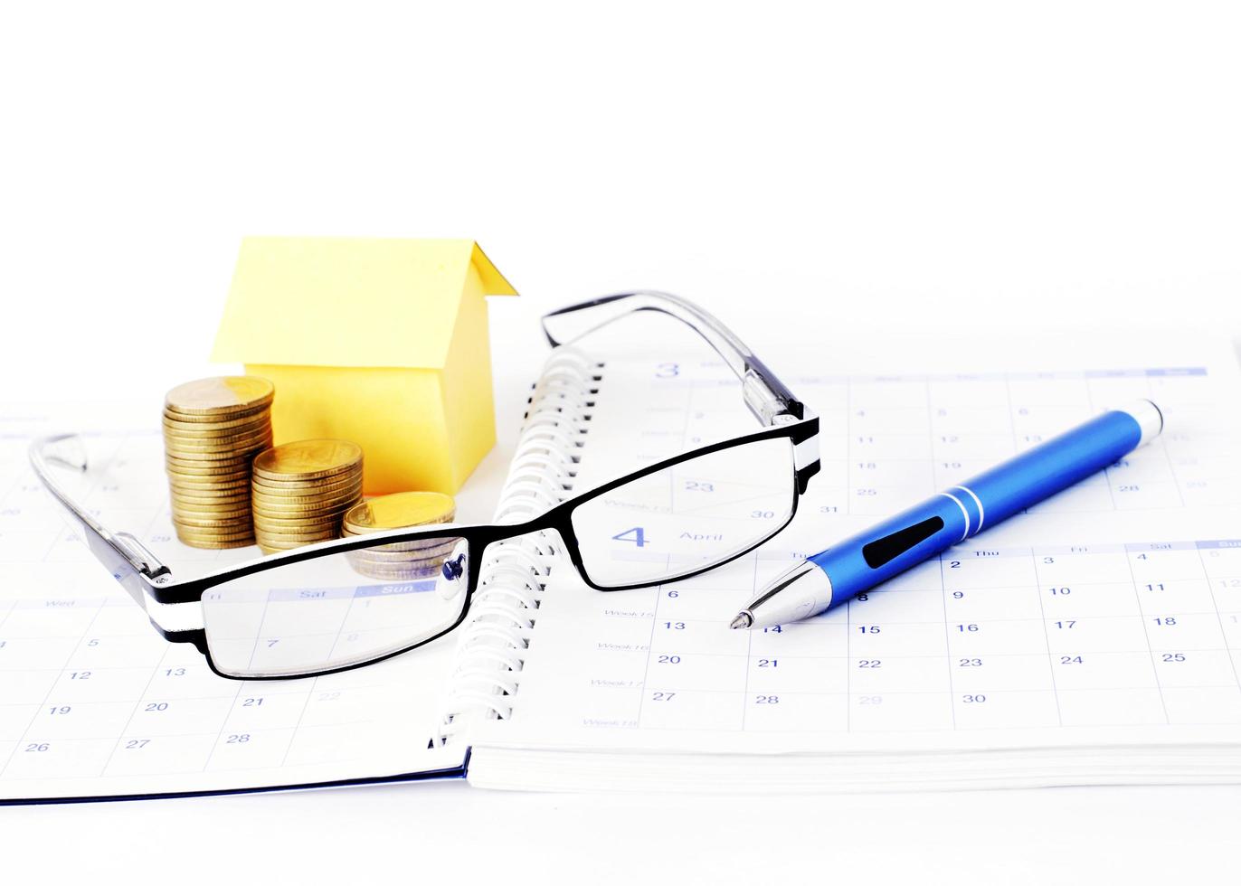 Loans concept with eyeglasses and blue pen with money coins and yellow paper house on calendar book page photo