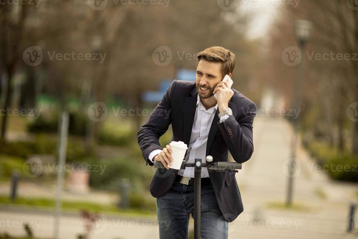 empresario casual montando un scooter mientras sostiene un café y habla por teléfono foto