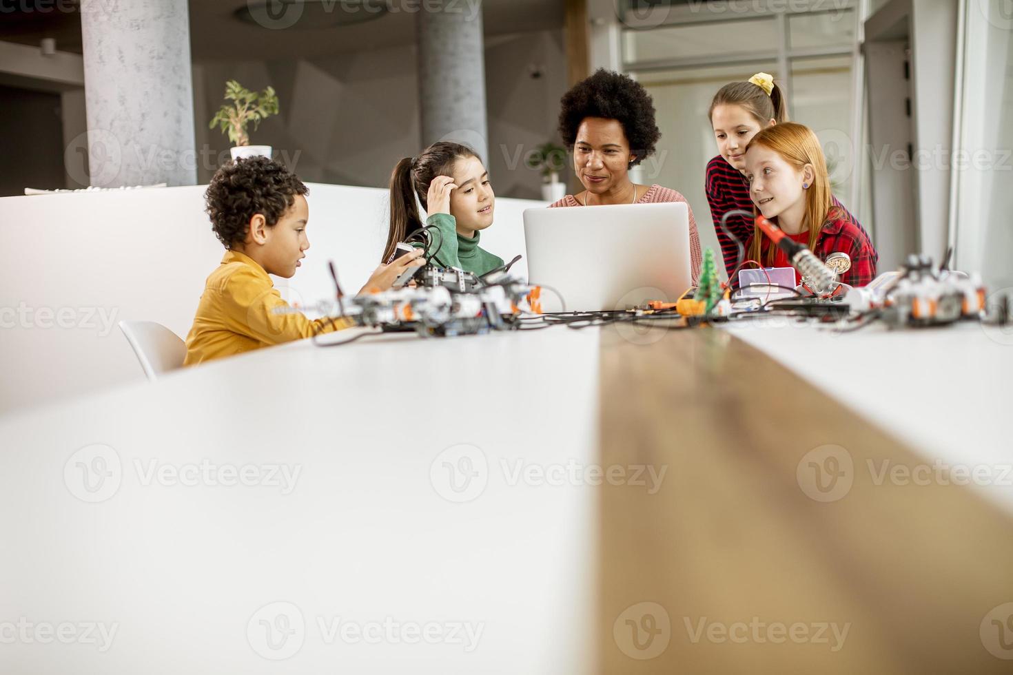 Niños felices con su profesora de ciencias afroamericana con programación de portátiles, juguetes eléctricos y robots en el aula de robótica foto