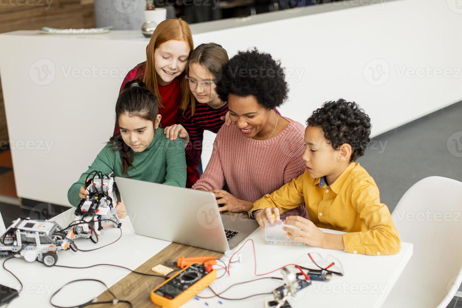 Niños felices con su profesora de ciencias afroamericana con programación de portátiles, juguetes eléctricos y robots en el aula de robótica foto