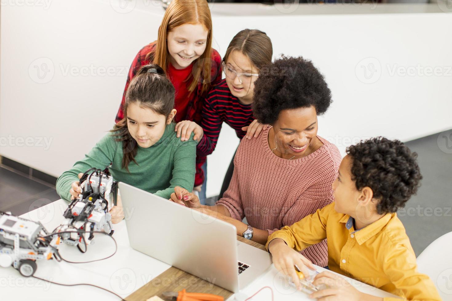 Niños felices con su profesora de ciencias afroamericana con programación de portátiles, juguetes eléctricos y robots en el aula de robótica foto