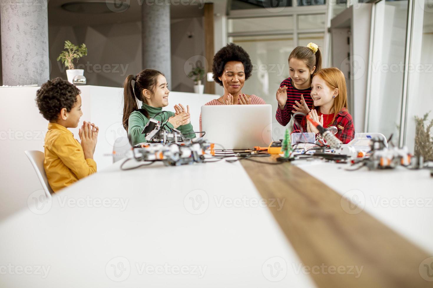 Niños felices con su profesora de ciencias afroamericana con programación de portátiles, juguetes eléctricos y robots en el aula de robótica foto