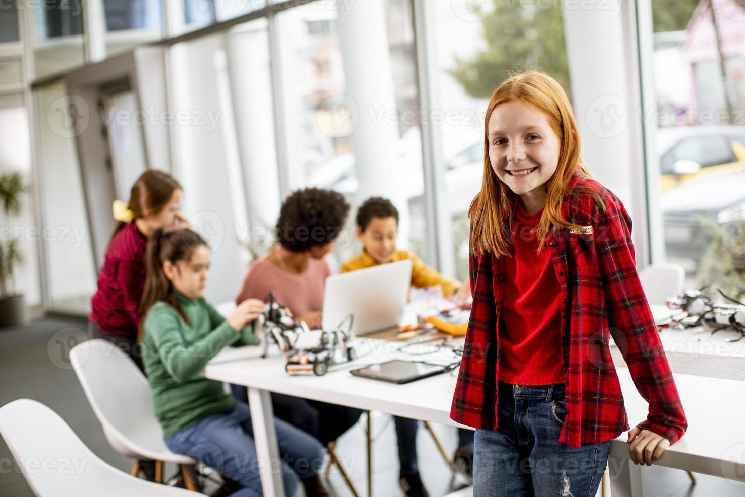 Niña linda de pie frente a los niños que programan juguetes eléctricos y robots en el aula de robótica foto