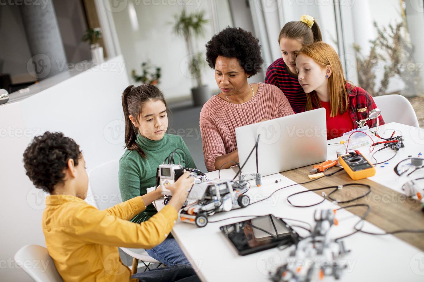 Happy kids with their African American female science teacher with laptop programming electric toys and robots at robotics classroom photo