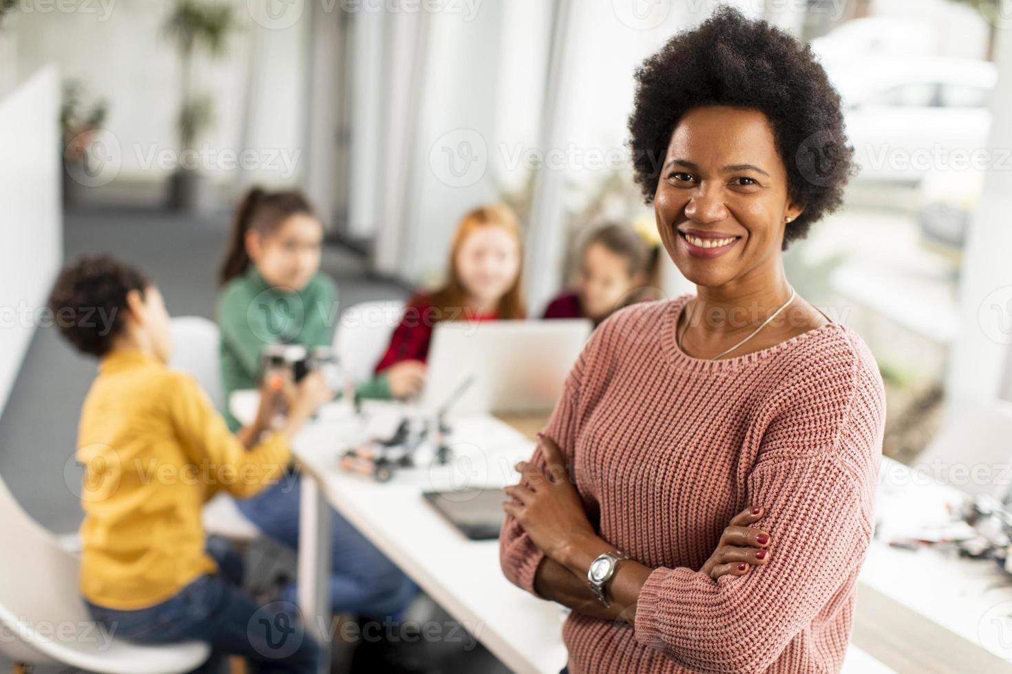 Profesora de ciencias afroamericana con un grupo de niños que programan juguetes eléctricos y robots en el aula de robótica foto