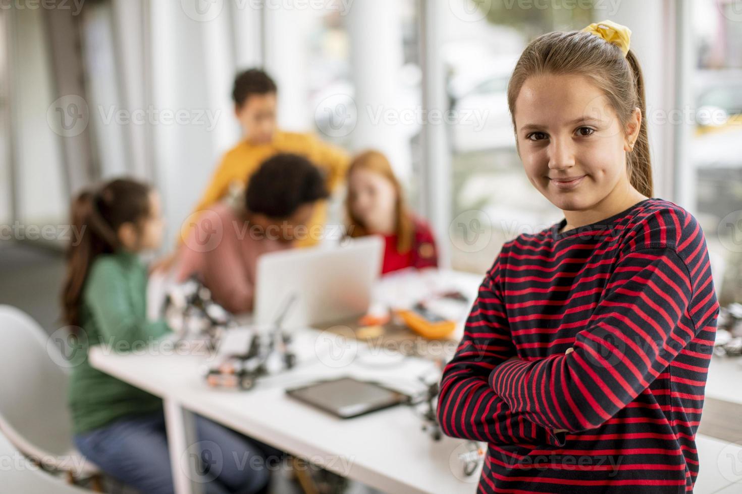 Niña linda de pie frente a los niños que programan juguetes eléctricos y robots en el aula de robótica foto