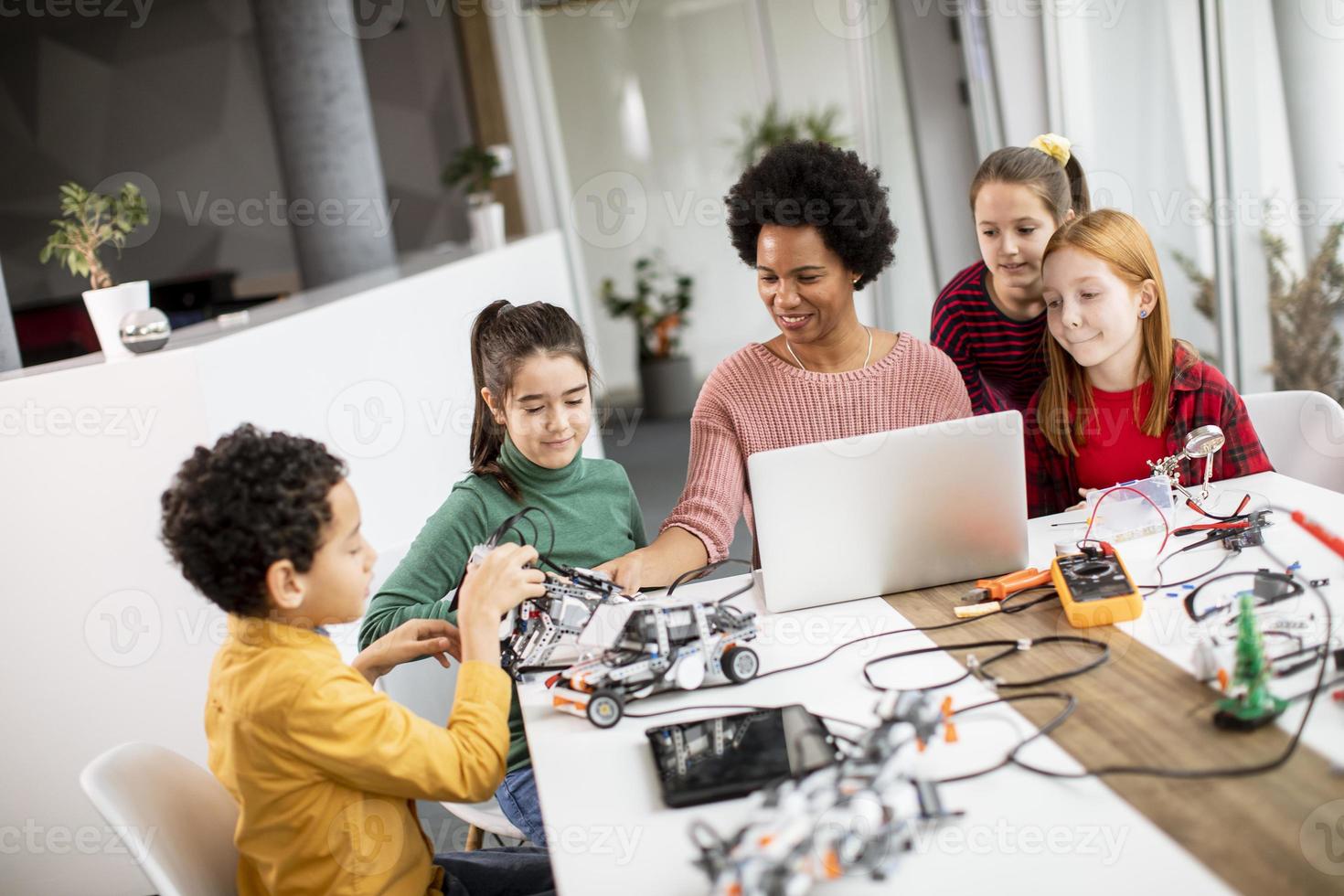 Niños felices con su profesora de ciencias afroamericana con programación de portátiles, juguetes eléctricos y robots en el aula de robótica foto