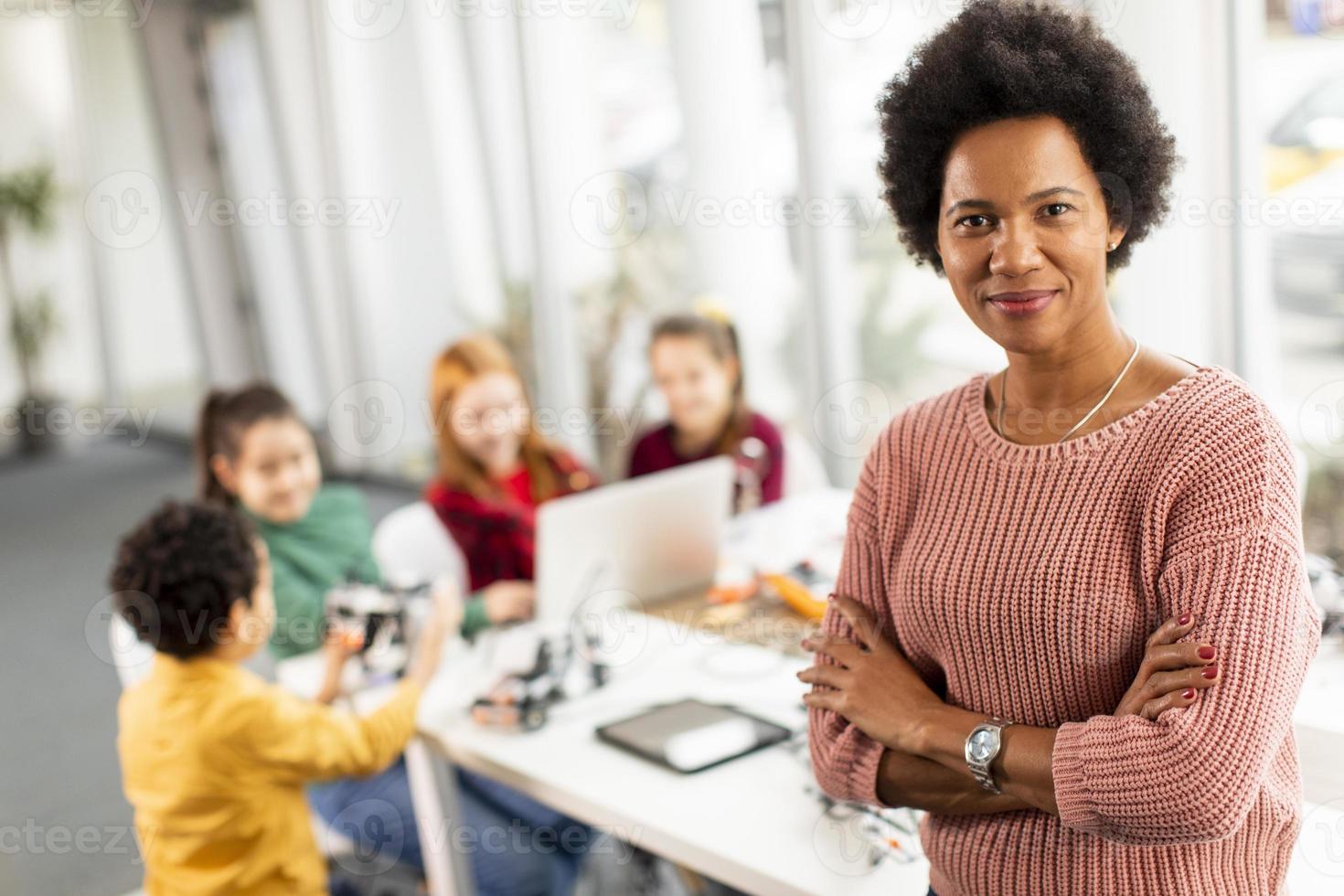 Profesora de ciencias afroamericana con un grupo de niños que programan juguetes eléctricos y robots en el aula de robótica foto