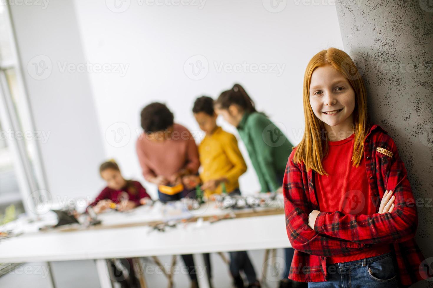 Niña linda de pie frente a los niños que programan juguetes eléctricos y robots en el aula de robótica foto