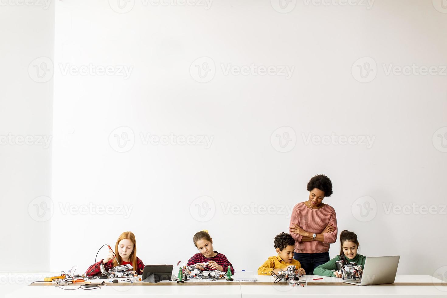 Niños felices con su profesora de ciencias afroamericana con programación de portátiles, juguetes eléctricos y robots en el aula de robótica foto