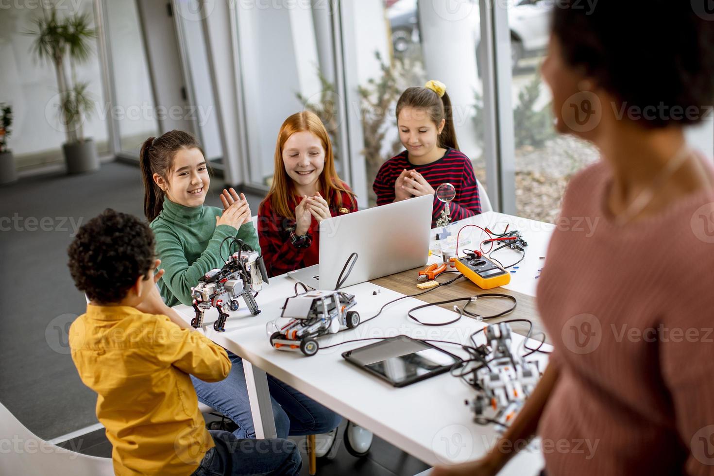 Happy kids with their African American female science teacher with laptop programming electric toys and robots at robotics classroom photo