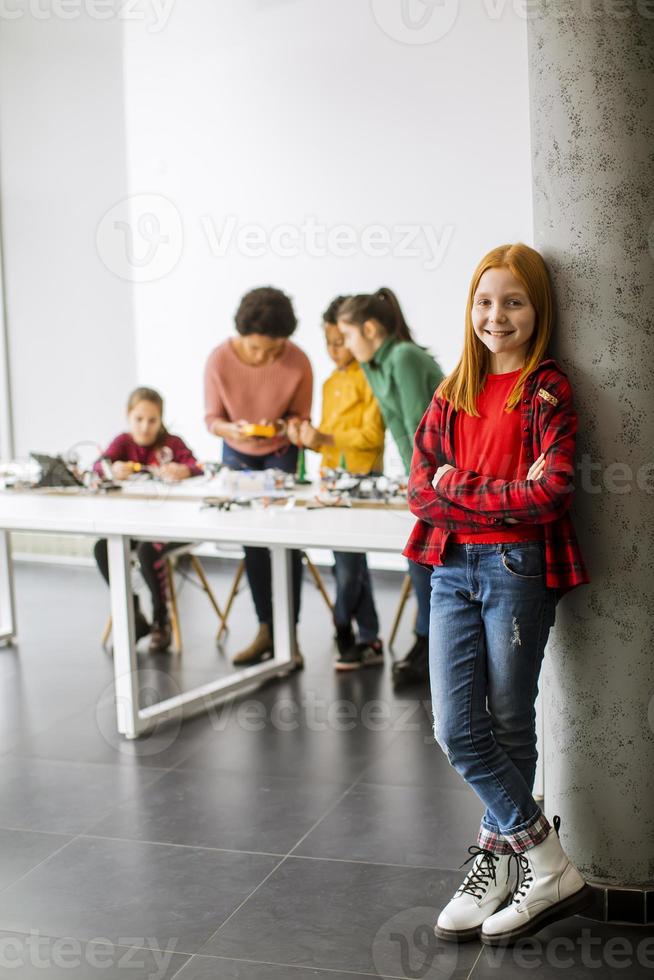 Niña linda de pie frente a los niños que programan juguetes eléctricos y robots en el aula de robótica foto