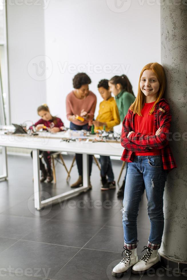 Niña linda de pie frente a los niños que programan juguetes eléctricos y robots en el aula de robótica foto