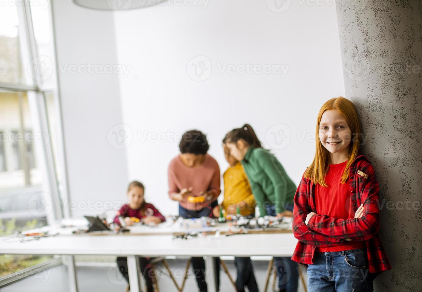 Niña linda de pie frente a los niños que programan juguetes eléctricos y robots en el aula de robótica foto