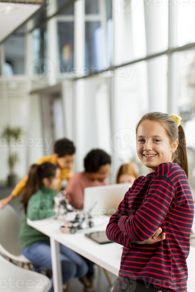 Niña linda de pie frente a los niños que programan juguetes eléctricos y robots en el aula de robótica foto