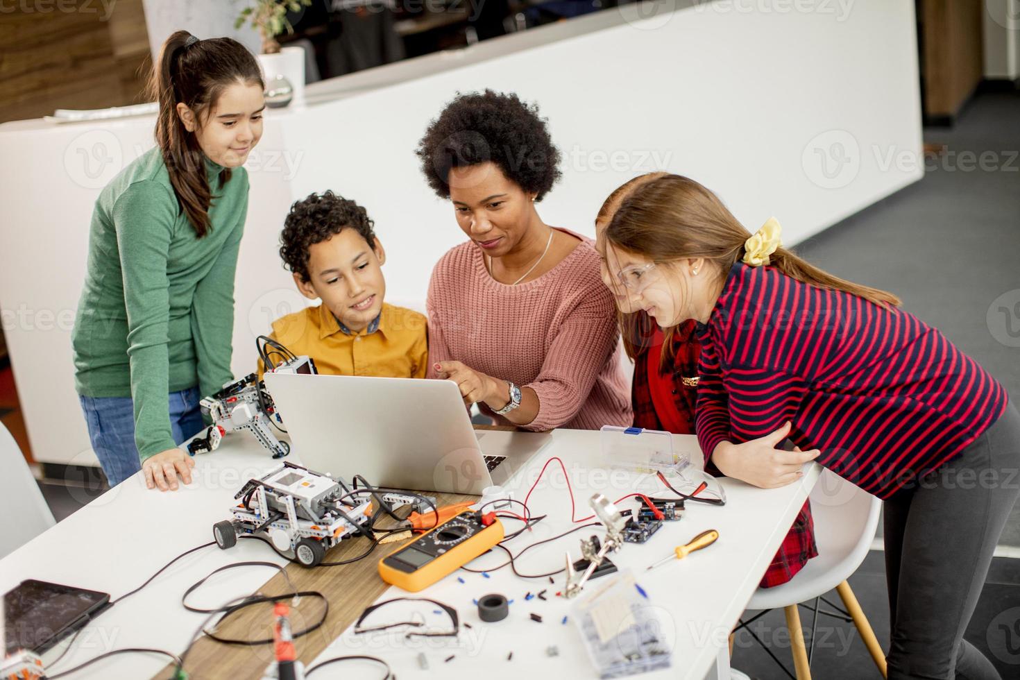 Niños felices con su profesora de ciencias afroamericana con programación de portátiles, juguetes eléctricos y robots en el aula de robótica foto