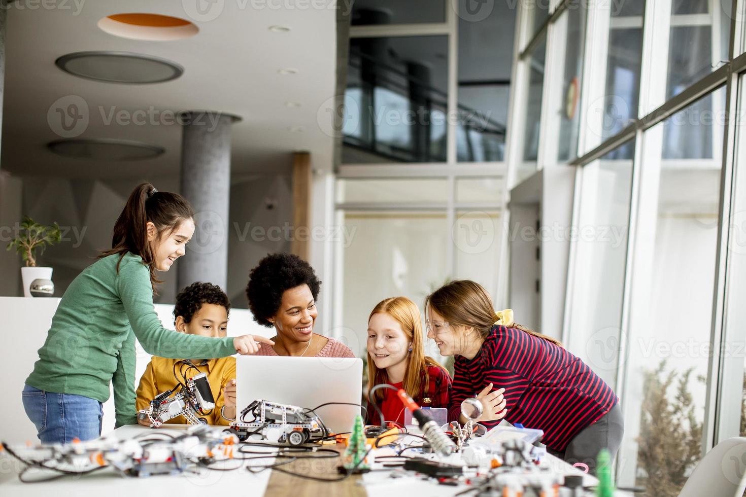 Niños felices con su profesora de ciencias afroamericana con programación de portátiles, juguetes eléctricos y robots en el aula de robótica foto