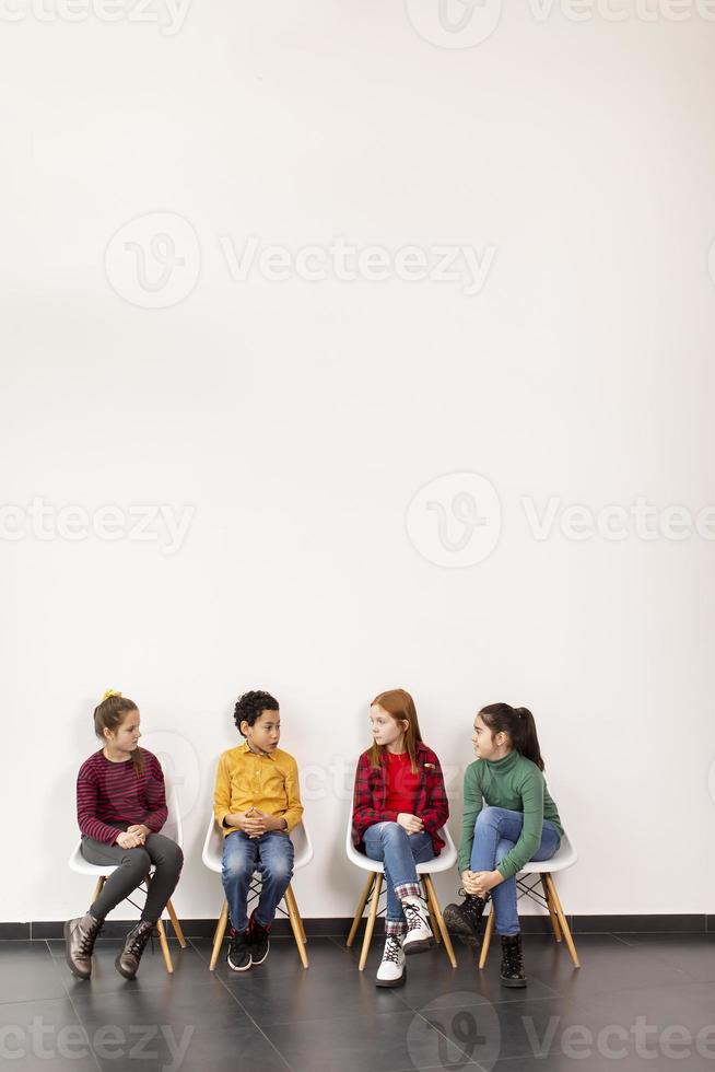 Retrato de lindos niños en jeans sentados en sillas contra la pared blanca foto