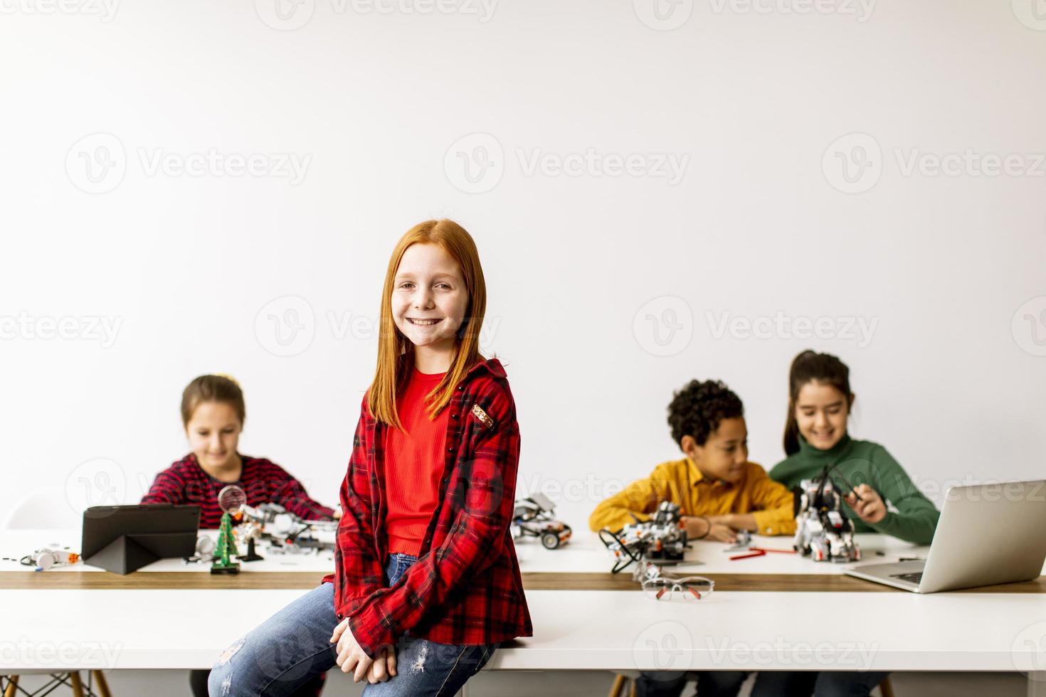 Niña linda de pie frente a los niños que programan juguetes eléctricos y robots en el aula de robótica foto