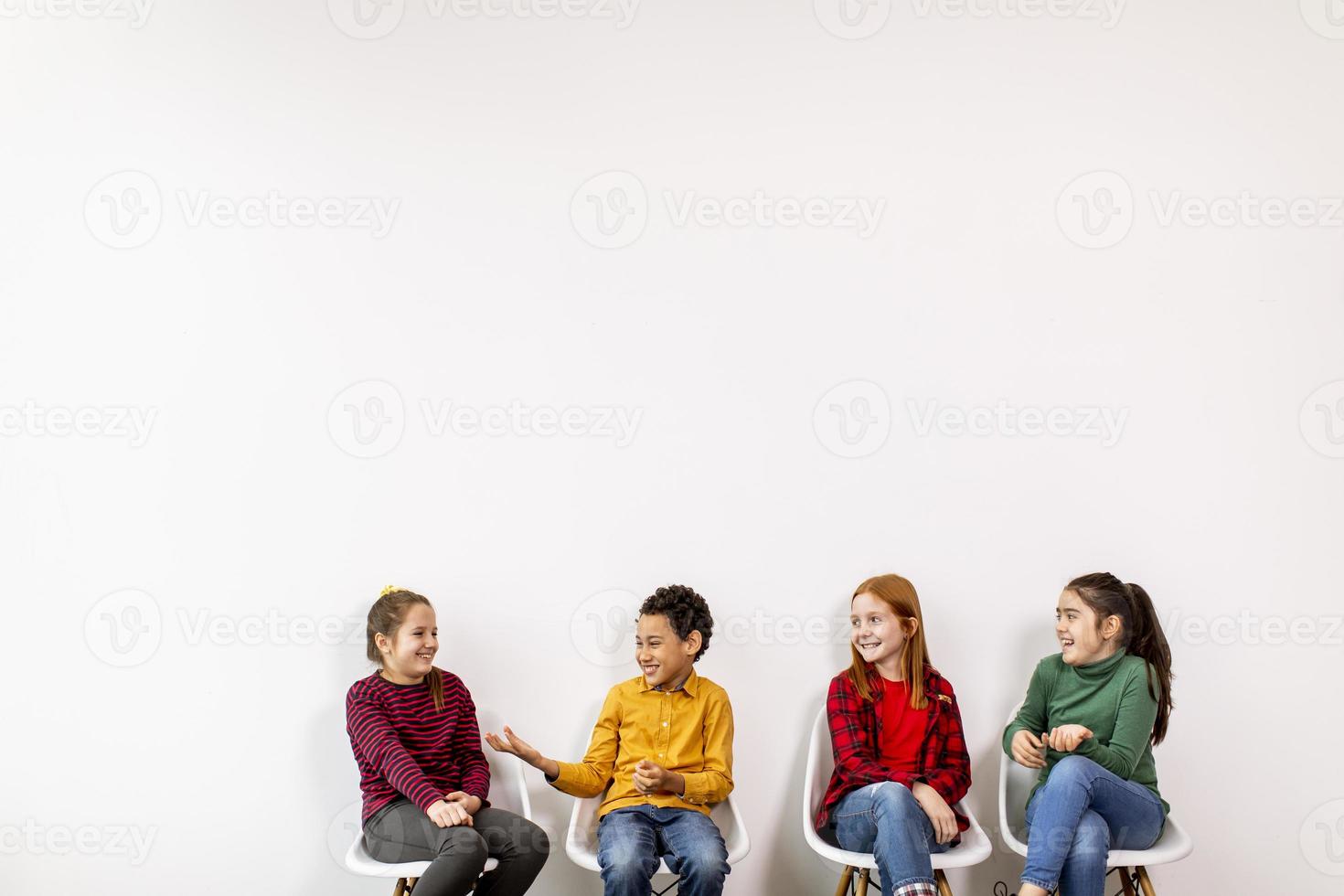 Retrato de lindos niños en jeans sentados en sillas contra la pared blanca foto