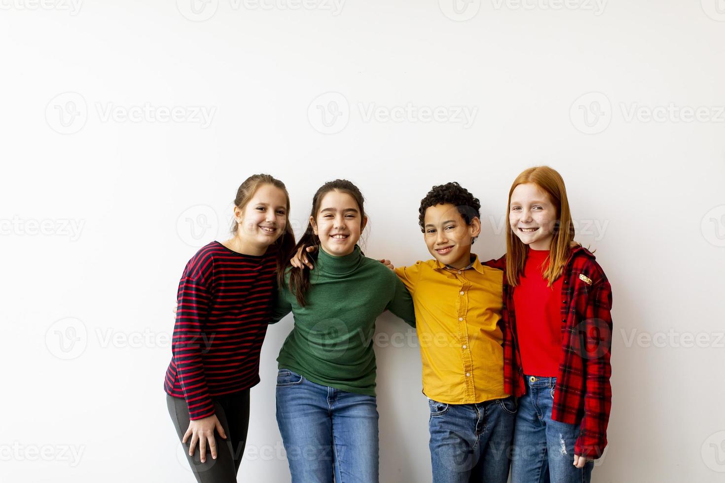 Retrato de lindos niños pequeños en jeans mirando a la cámara y sonriendo y de pie contra la pared blanca foto