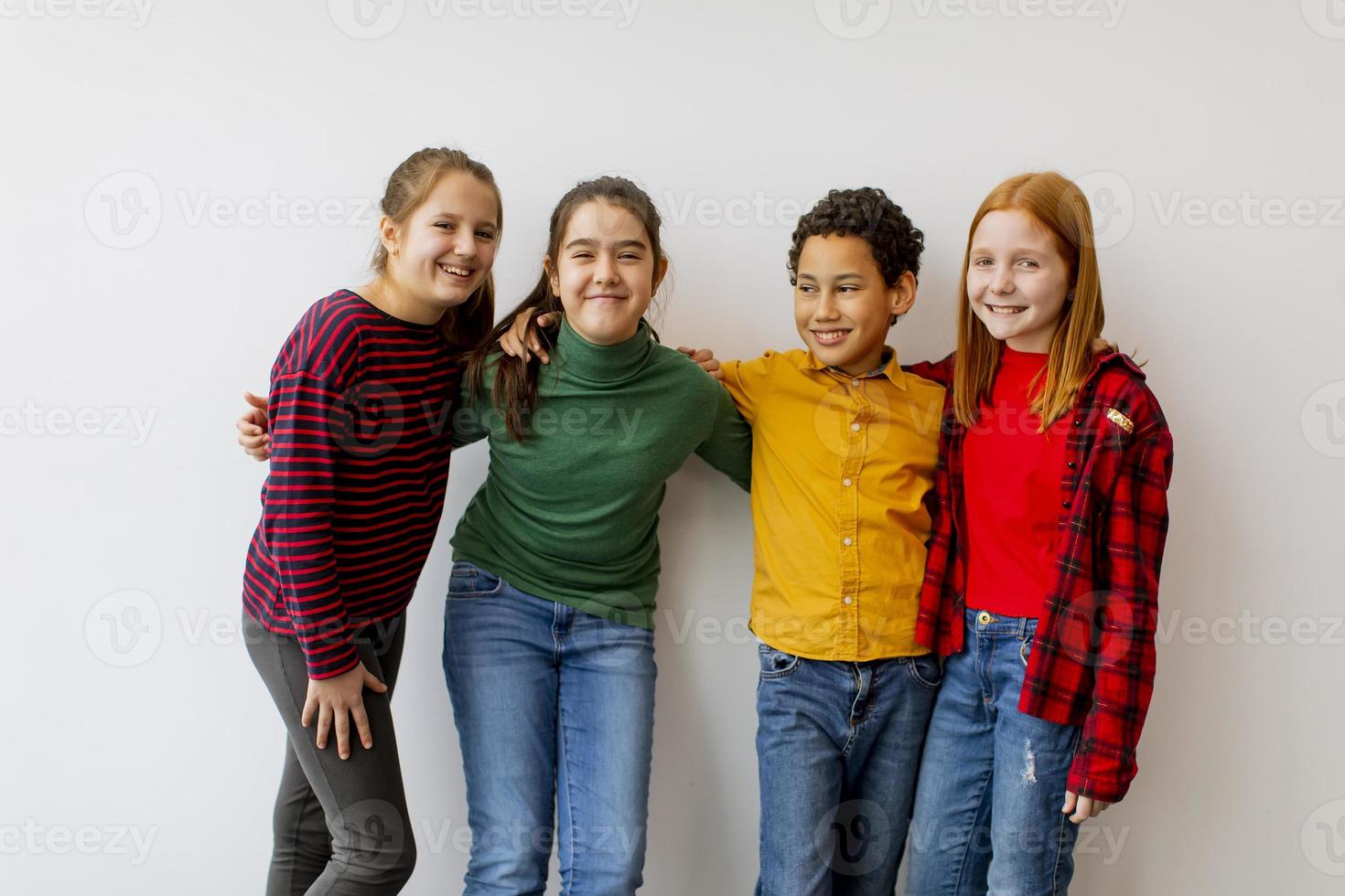 Retrato de lindos niños pequeños en jeans mirando a la cámara y sonriendo y de pie contra la pared blanca foto