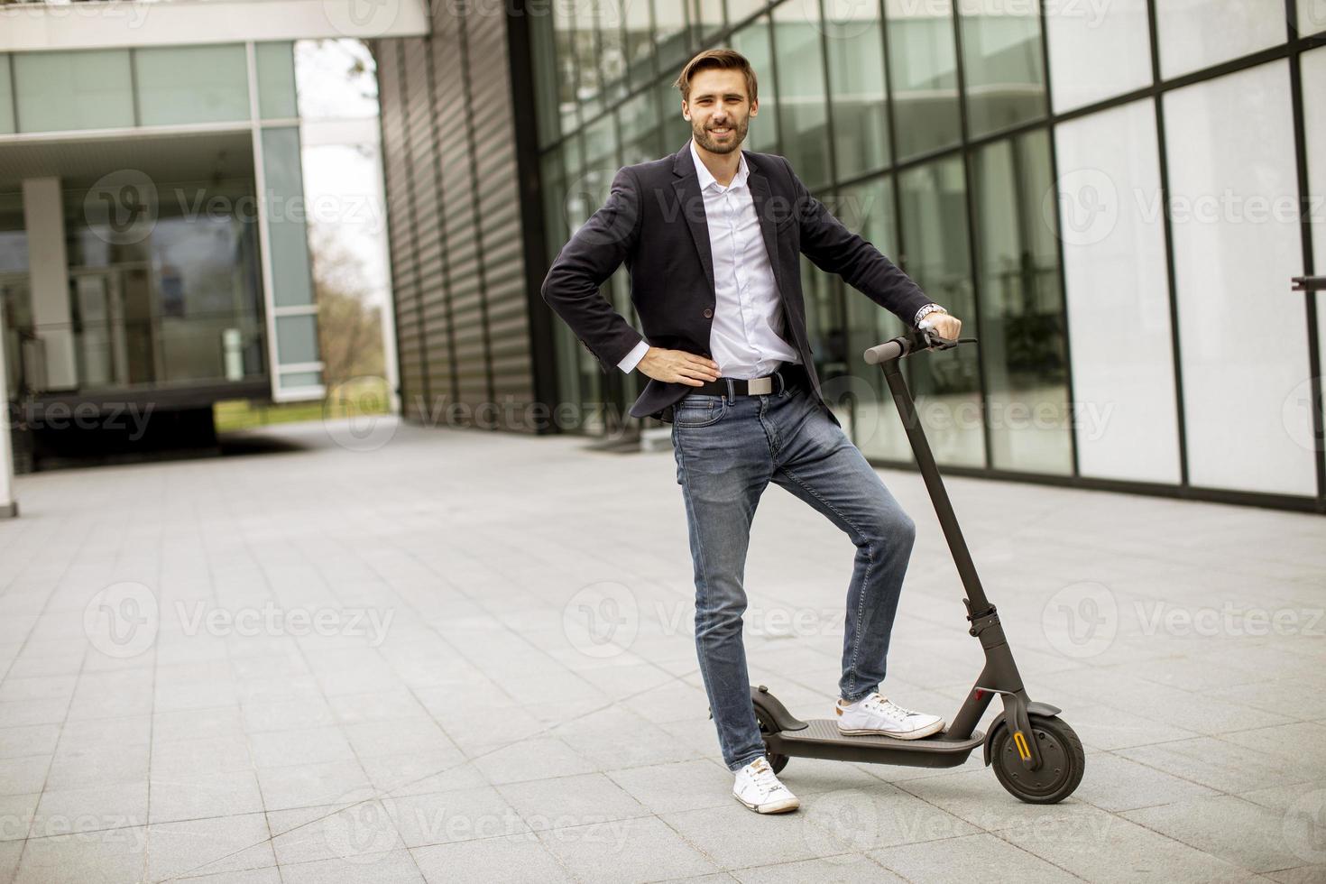 Hombre de negocios joven en ropa casual de pie en scooter eléctrico por un edificio de oficinas en una reunión de negocios foto