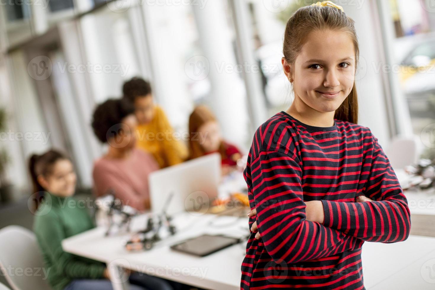 Niña linda de pie frente a los niños que programan juguetes eléctricos y robots en el aula de robótica foto