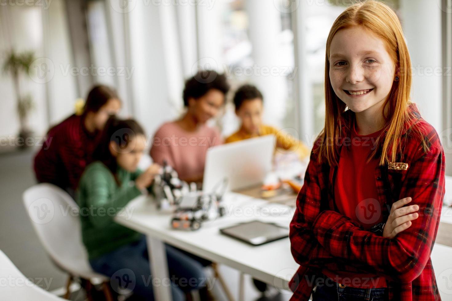 Cute little girl standing in front of kids programming electric toys and robots at robotics classroom photo