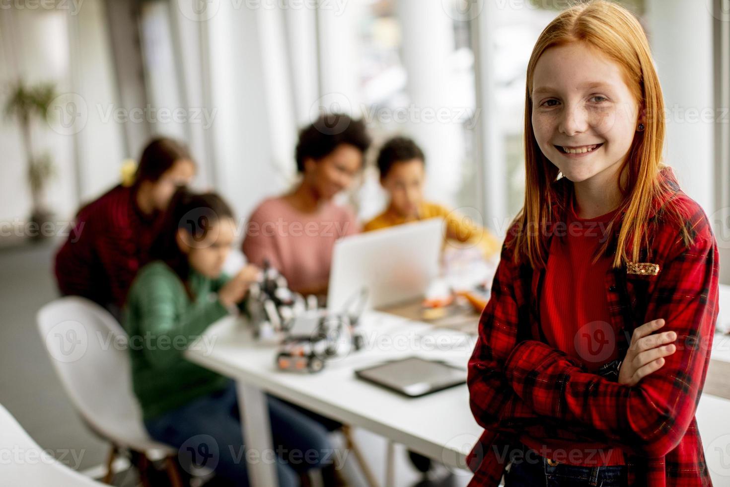 Niña linda de pie frente a los niños que programan juguetes eléctricos y robots en el aula de robótica foto