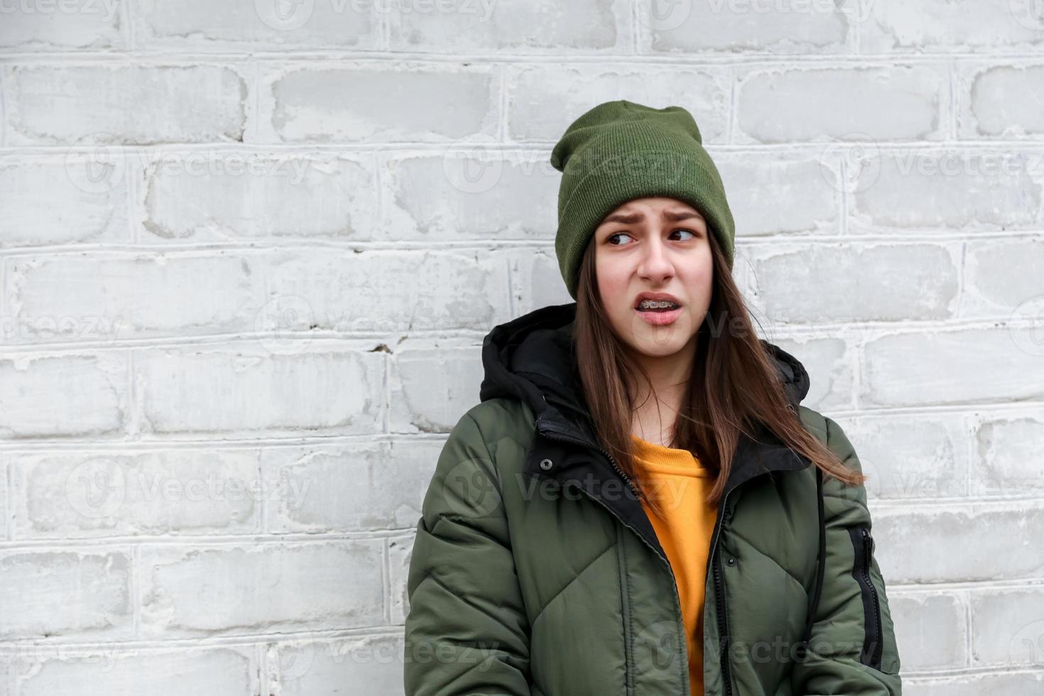 Portrait of a beautiful incredulous girl with braces in a yellow sweater and khaki hat that stands near a white brick wall photo