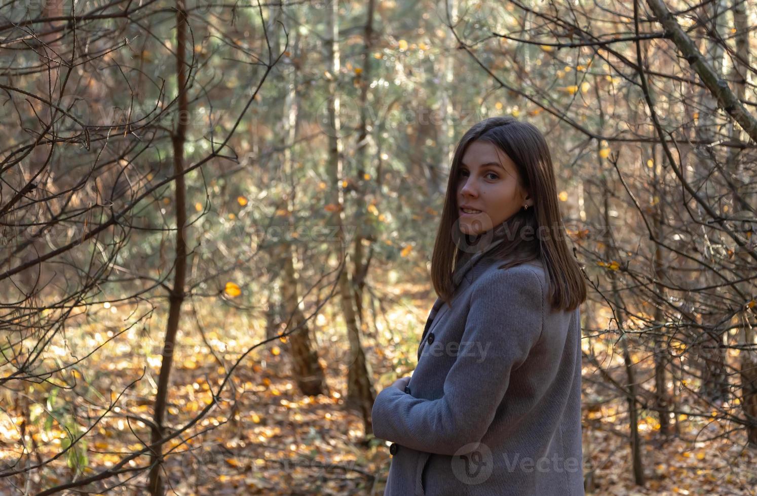 Chica medio vuelta con un abrigo gris que se encuentra en el bosque en un soleado día de otoño foto