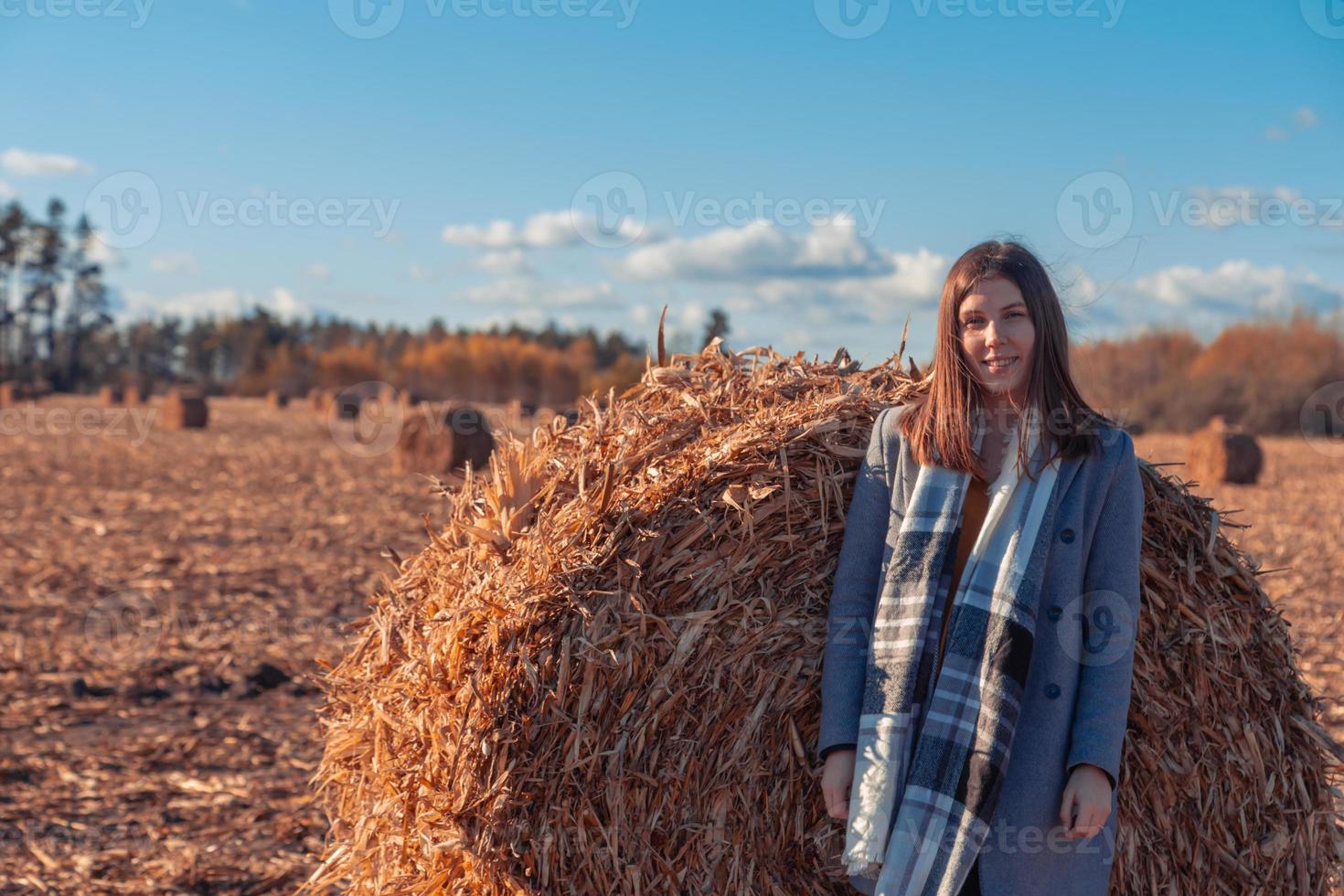 A girl of European appearance in a gray coat stands in a field near a larger bale with hay against a blue sky photo