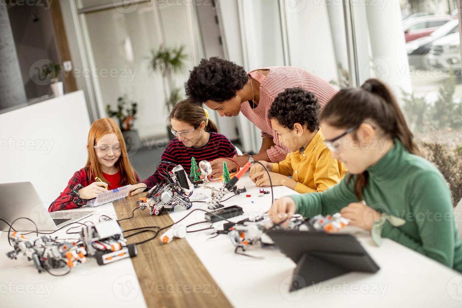 Niños felices con su profesora de ciencias afroamericana con programación de portátiles, juguetes eléctricos y robots en el aula de robótica foto