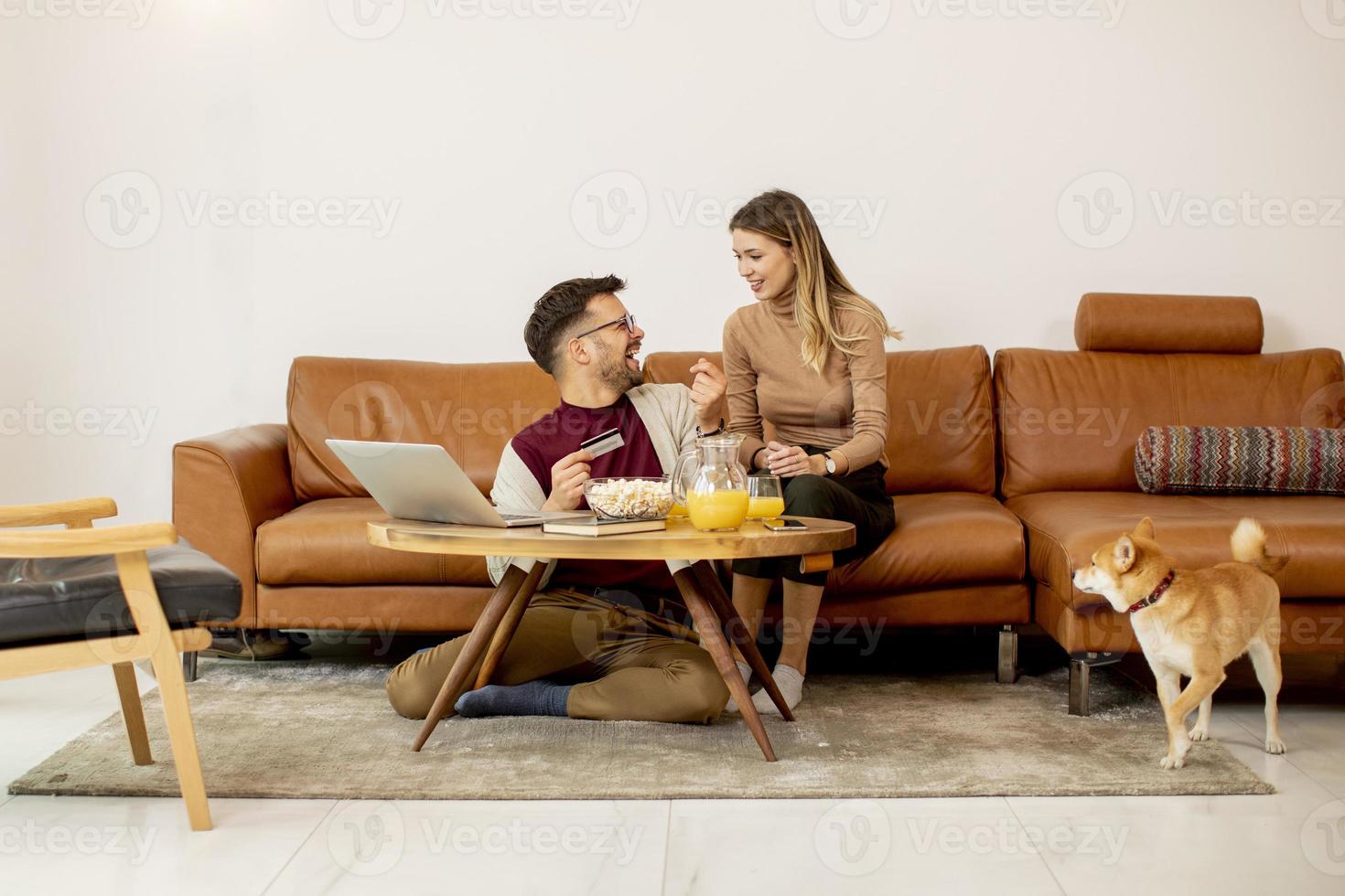 Young woman and young man using laptop for onlin payment while sitting by sofa with their shiba inu dog at home photo