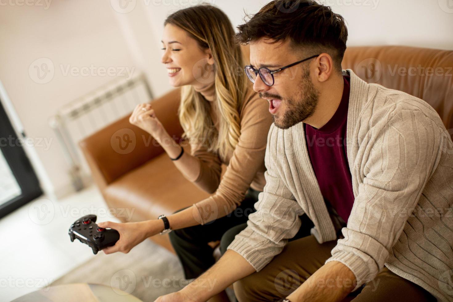 Pareja joven jugando videojuegos en casa sentado en el sofá y divirtiéndose foto