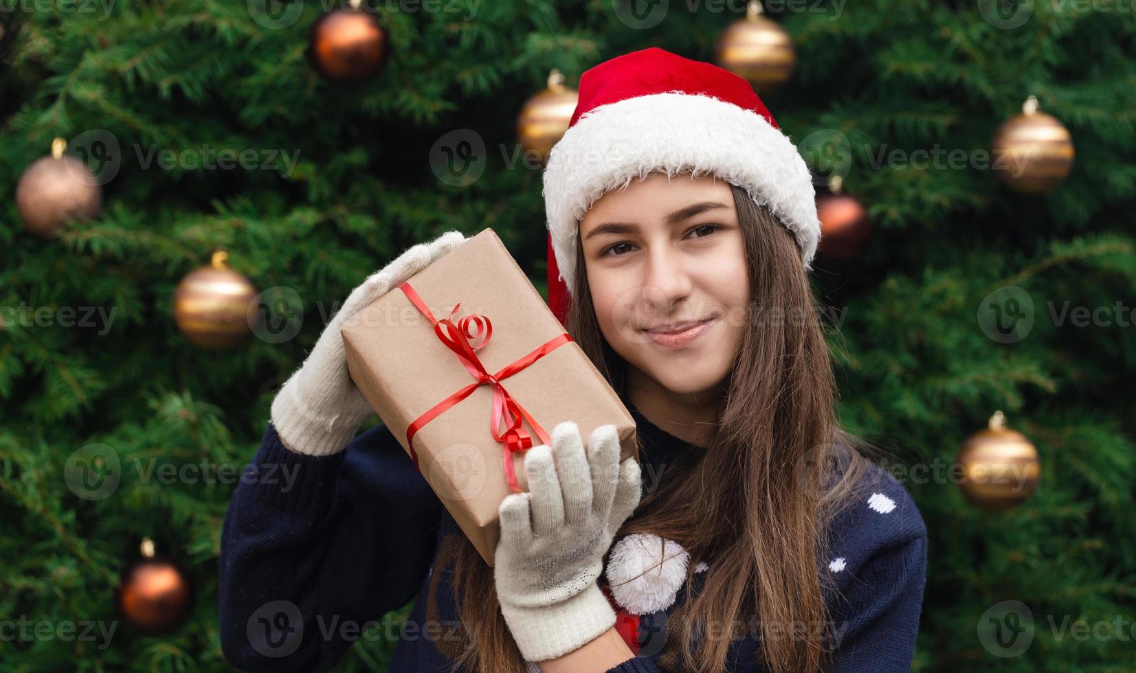 A young girl wearing a santa claus hat gives a gift made of craft paper with a red ribbon photo