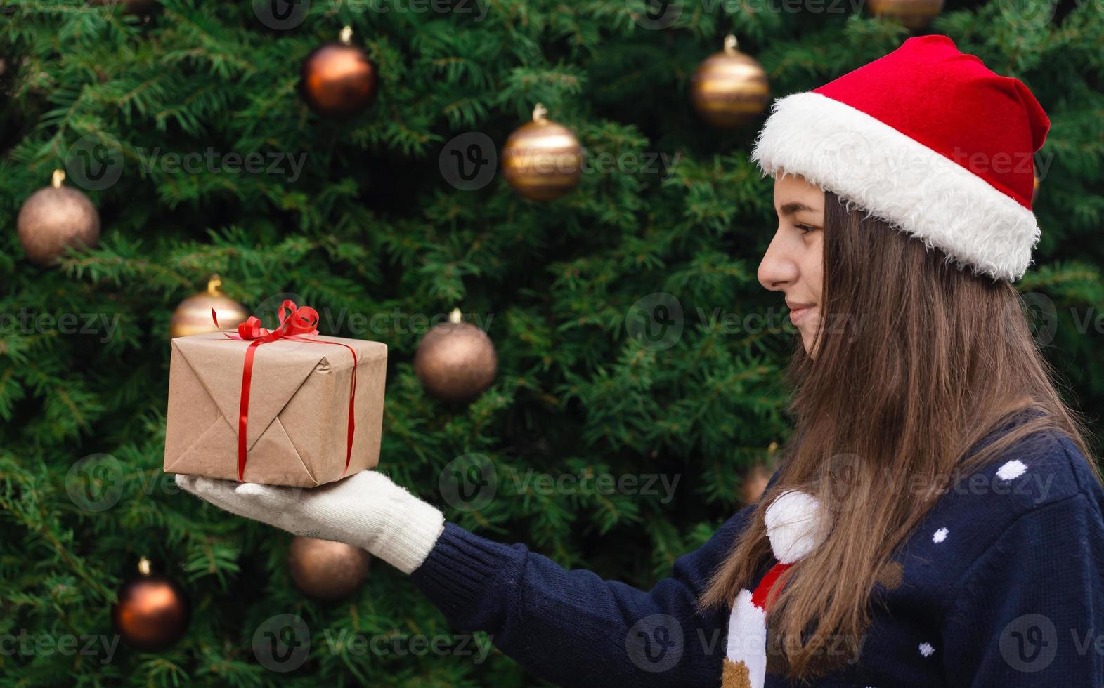 A young girl wearing a santa claus hat gives a gift made of craft paper with a red ribbon photo