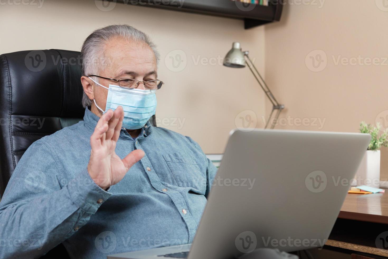 Hombre senior en mascarilla trabajando o comunicándose en un portátil en casa foto