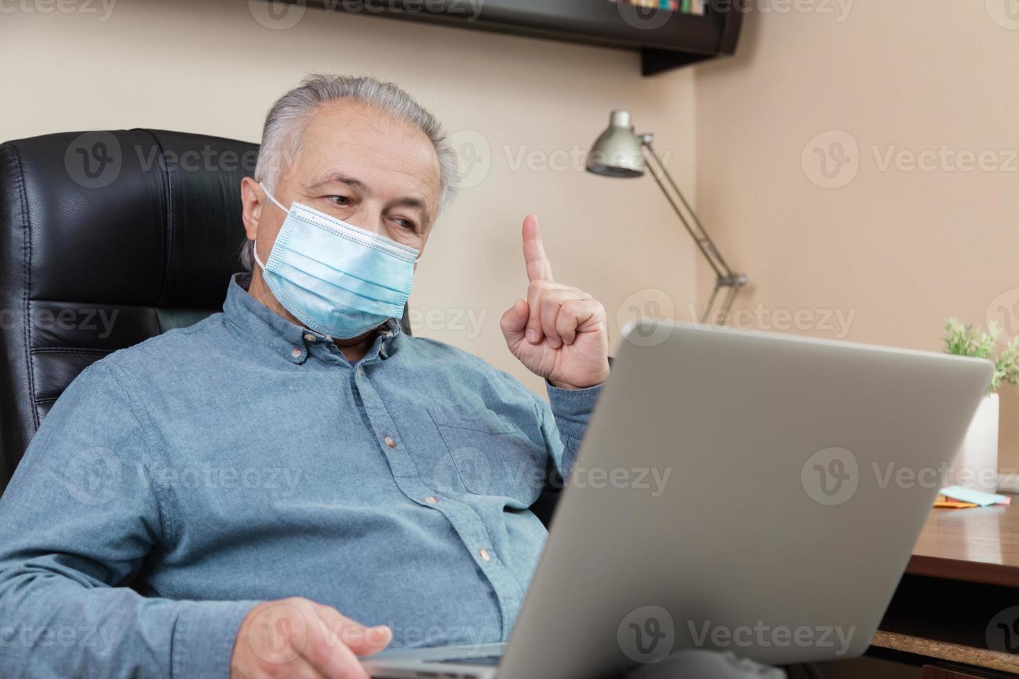 Hombre senior en mascarilla trabajando o comunicándose en un portátil en casa foto