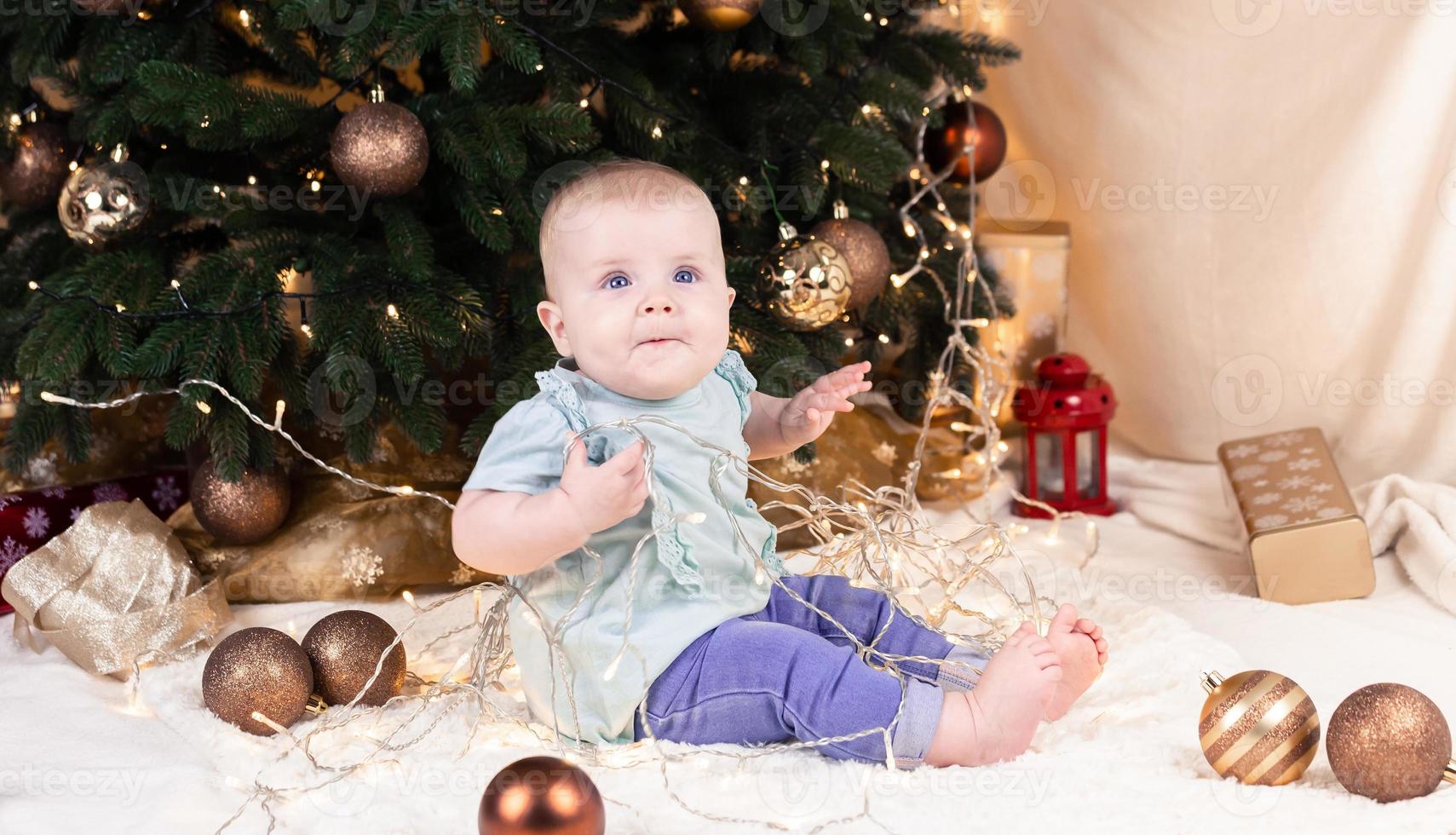 un bebé en jeans se sienta cerca de un árbol de navidad y juega con una guirnalda en la que está enredado foto