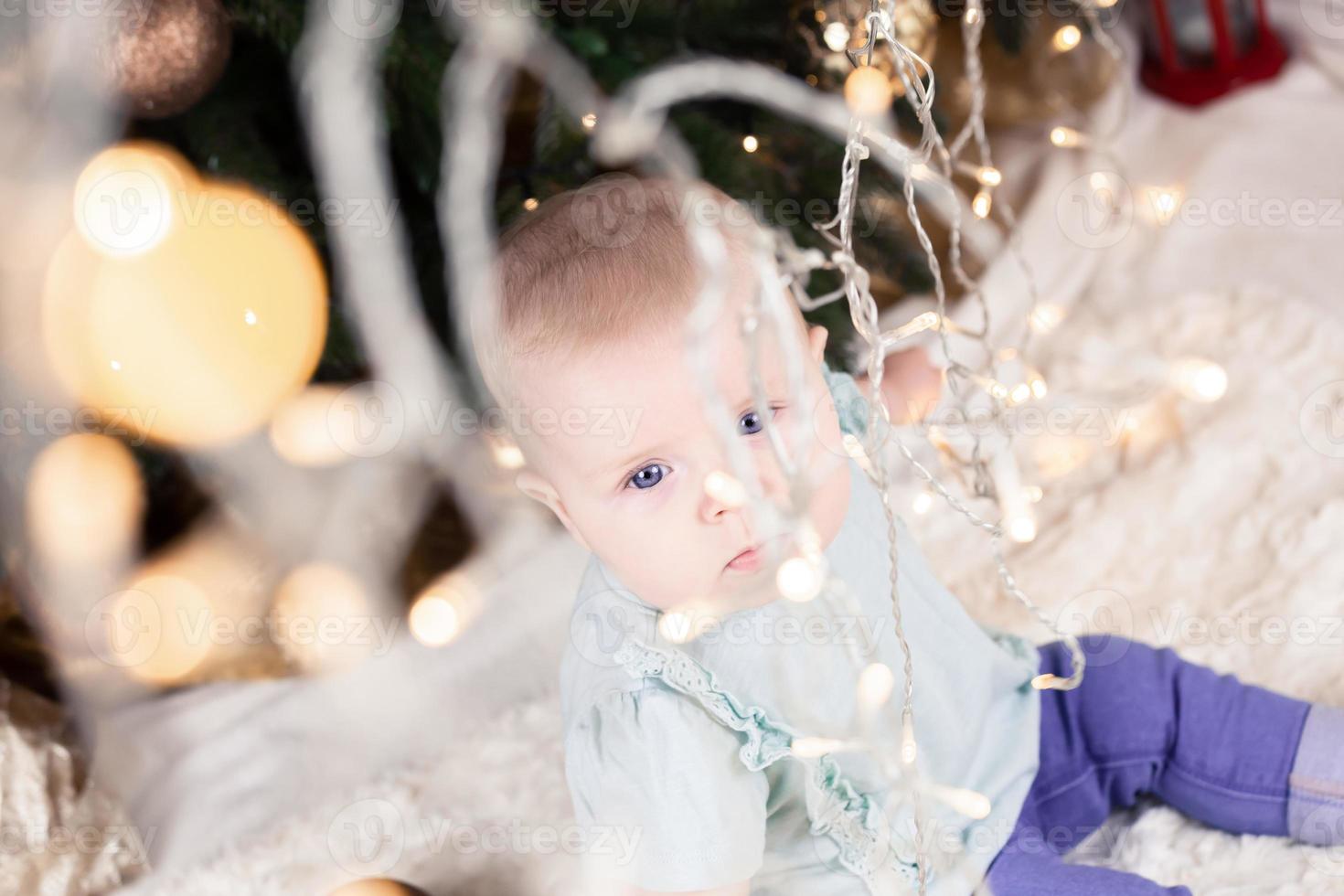 bebé en jeans se sienta cerca de un árbol de navidad y mira a través de una guirnalda foto