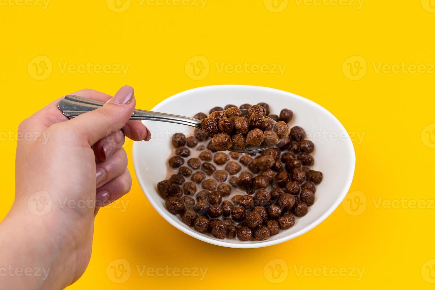 Mujer mano sosteniendo una cuchara en un tazón blanco con bolas de cereal de maíz de chocolate y leche sobre fondo amarillo foto