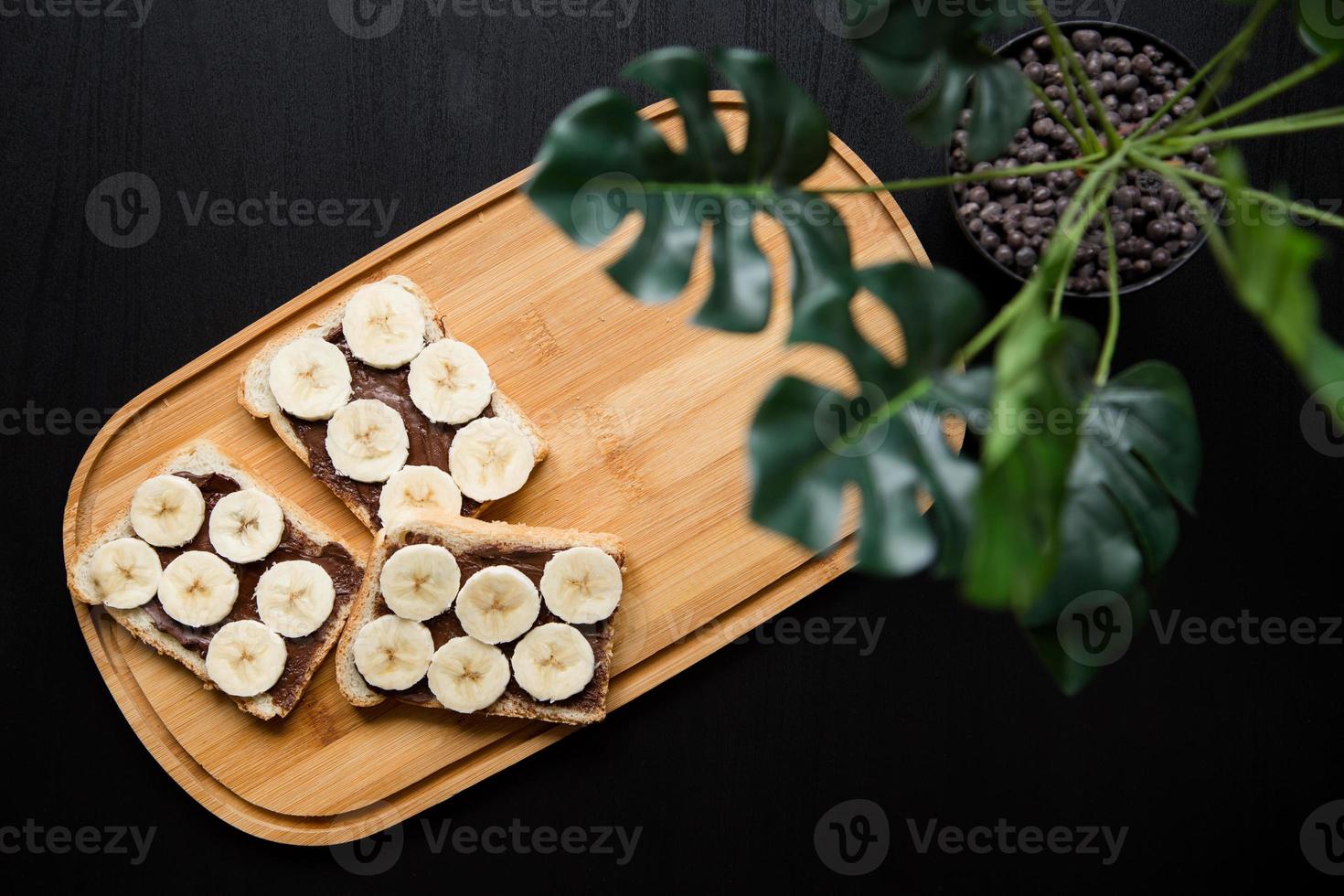 Three banana white bread toasts spread with chocolate butter on a chopping board with leaves on a dark background photo