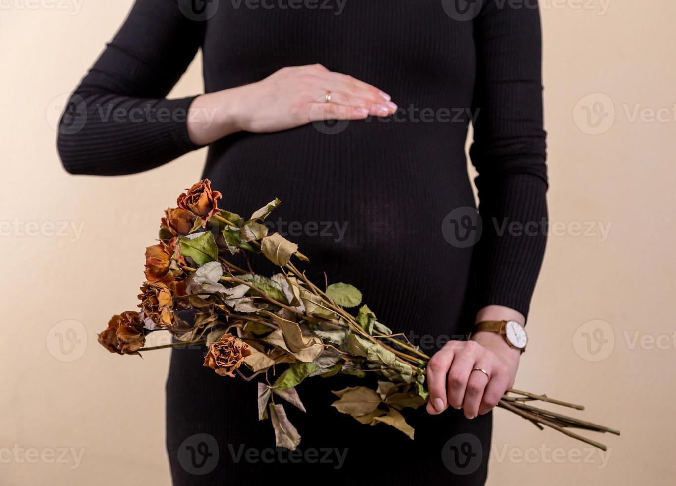 Vista lateral de una atractiva mujer embarazada acariciando su vientre y sosteniendo un ramo de rosas secas foto
