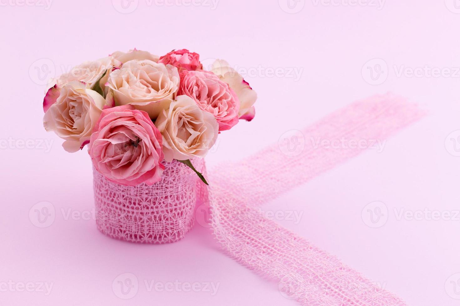 A bouquet of beautiful roses stands in a small bucket on a lace ribbon on a pink background with space for text photo